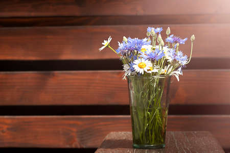 A bouquet of daisies and cornflowers in a glass vase on a wooden table against the background of a wooden wall. Place for an inscription. Selective focus. Copyspace.の写真素材