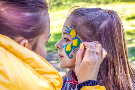 An artists hand draws a blue-yellow flower on the face of a little girl. A child with a funny face...