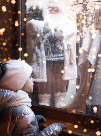 Girl looking out the window at Christmas decorations in a store. Evening Christmas shopping lightsの写真素材
