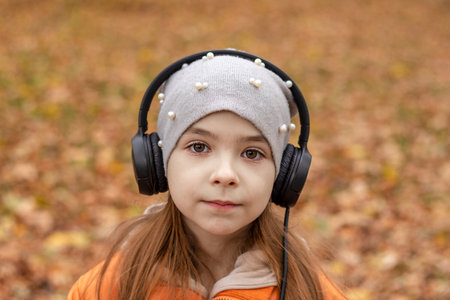 A girl listens to music on headphones in an autumn park and looks at the camera.の写真素材