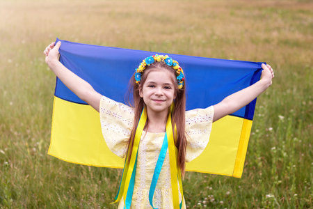 A girl in a wreath of yellow and blue flowers holds the flag of Ukraine in her hands against the background of a field.の写真素材