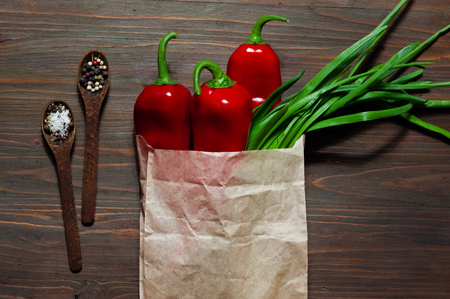 Red pepper, green onion in a paper bag and wooden spoon with spice on a dark wooden surface, space...