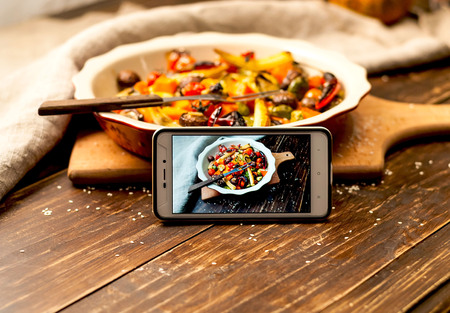 Spicy hot vegetables, cooked on an grill in ceramic bowl on wooden background with smartphone. Food photo for social networks, copy space, closeupの写真素材