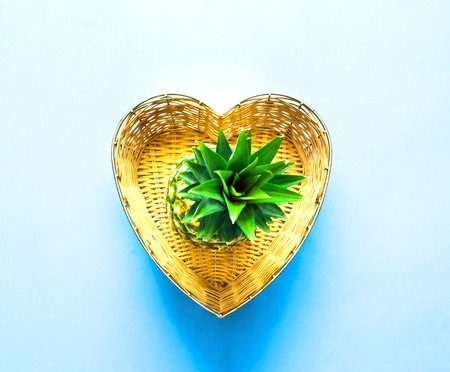 Pineapple in wicker basket in the shape of heart on blue background. Tropical fruit concept. Close-up, copy space.の写真素材