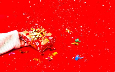 Female hand holds Christmas gift in festive packaging on red background with confetti. Copy space, close-upの写真素材