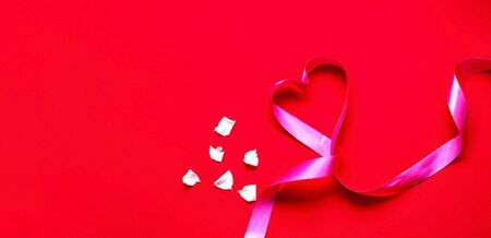Festive pink ribbon in the shape of heart on red background with white rose petals. Mothers Day concept. Template mock up of greeting card or text design. Close-up, copy spaceの写真素材