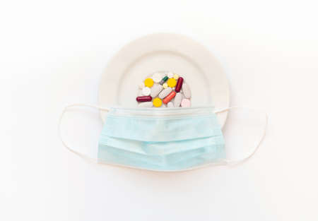 Medical protective mask and multicolored pills in white plate on isolated background. Medicine for...