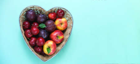Fruit for the Jewish New Year. Red apples and plums in straw basket made in the shape of heart on green background. Copy spaceの写真素材
