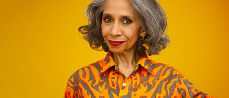 An elegant woman with gray hair and a bright orange patterned shirt stands against a yellow background, exuding confidence and styleの素材