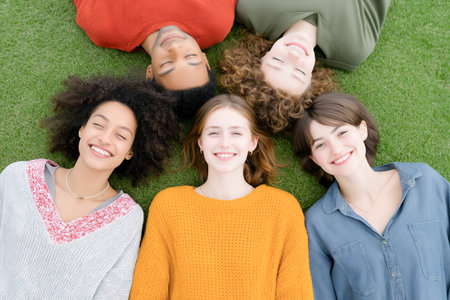 Diverse group of young adults lies on green grass, smiling and enjoying each other's company, capturing the essence of friendship and happiness in a natural environmentの素材