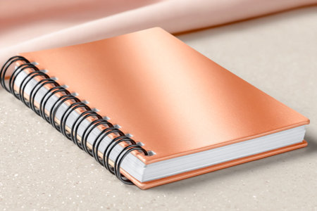Copper notebook with spiral binding lies on a smooth surface, accompanied by soft fabric, creating an inviting atmosphere for writing and creativityの素材