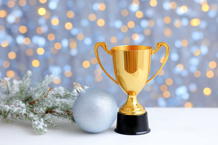 Golden trophy stands next to a silver ornament and evergreen branches, with a backdrop of blurred festive lights, creating a celebratory atmosphere for holidaysの素材