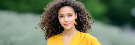 Confident young woman with curly hair in a yellow dress poses outdoors, surrounded by lush greenery, radiating beauty and a vibrant fashion senseの素材