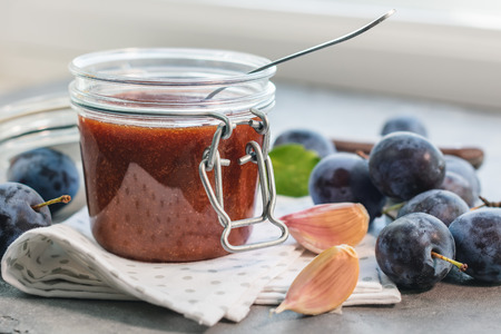 Plum sauce in a jar, garlic and blue ripe plums on a table.