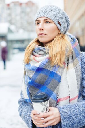 Girl in grey coat, hat and blanket scarf with cup of coffee on winter street, winter street style...
