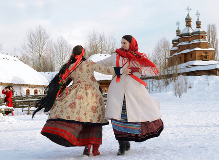 Two girls in national costumes dancing on the square in front of a wooden church on the snow during Traditional Ukrainian Christmas celebration in Kiev Historical Park in 2013 year.のeditorial素材