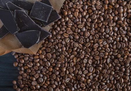 Roasted fragrant beans of black coffee are scattered on a black wooden table, on which black chocolate is lyingの写真素材