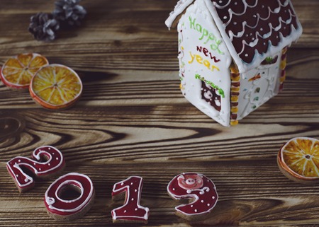 Christmas card on wood background gingerbread red numbers 2019 with slices of orange and white gingerbread house with a brown roof, a window and the inscription happy New Year on a white wallの写真素材
