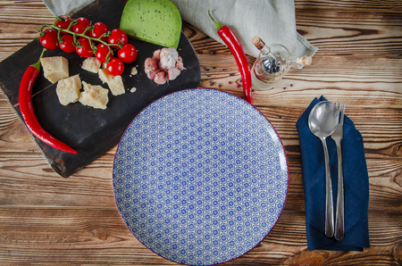 Parmesan, cherry tomatoes, chilli peppers, green cheese and garlic lie on a dark board that stands on a wooden table next to a beautiful plateの写真素材