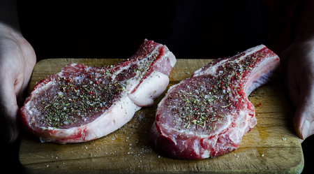 hands are holding a board on which are two raw pork steaks sprinkled with spices and ready to cookの写真素材
