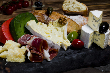 Camembert, brie, parmesan cheese, green cheese with basil, red chilli cheese and meat lie on a dark board next to tomatoes, olives, garlic, nuts, honey, a glass of white wine and bread on a wooden tableの写真素材