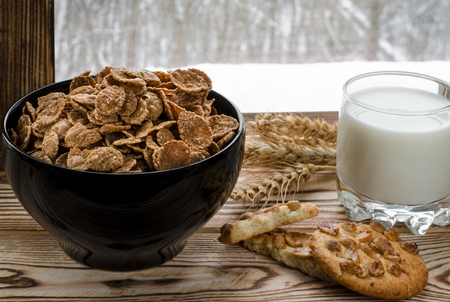 Cereal flakes lie in a black plate, which stands on a wooden table next to a glass of milk, biscuits and ears of wheat against the background of a winter forestの写真素材