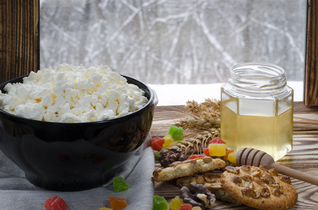 cottage cheese lie in a black plate, which stands on a wooden table next to a honey, biscuits, nuts, colorful candied fruits and ears of wheat against the background of a winter forestの写真素材