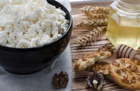 cottage cheese lie in a black plate, which stands on a wooden table next to a honey, biscuits, nuts, colorful candied fruits and ears of wheat against the background of a winter forestの写真素材