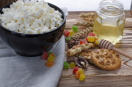 cottage cheese lie in a black plate, which stands on a wooden table next to a honey, biscuits, nuts, colorful candied fruits and ears of wheat against the background of a winter forestの写真素材