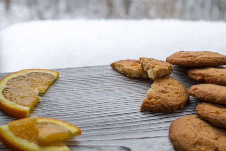 Round orange biscuits with colorful candied fruits and a slice of juicy orange lying on a wooden table against the background of winter snow-covered forest.の写真素材