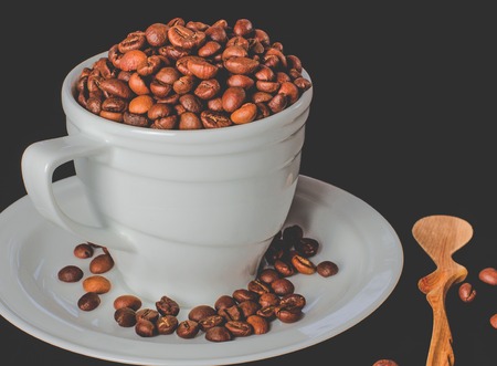 A white cup full of coffee beans stands on a white saucer, which stands on a black background next to a wooden spoonの写真素材
