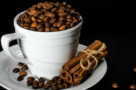 A white cup full of coffee beans stands on a white saucer, which stands on a black background next to a wooden spoonの写真素材