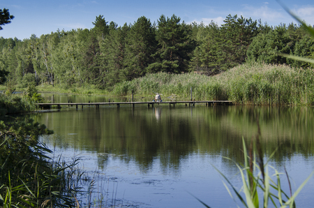 A wooden bridge over a river in the forest where a fisherman sits on a background of green forest and blue skyの写真素材