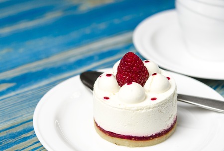 White airy cake with raspberry layer and raspberry berry lies on a white round plate next to a white cup, which stands on a blue wooden tableの写真素材
