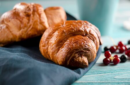 Fresh ruddy croissants with berries lie on a wooden table next to fresh black currant berries, red currants, cherries and a cup of coffeeの写真素材