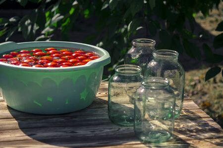 A fresh crop of red tomatoes washed in clear water, floats in a round plastic bowl, which stands on a wooden table, next to glass jars in the garden. Ready for winter conservationの写真素材