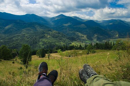The legs of two people who lie on the grass against the backdrop of high mountain ranges. Climbing to the topの写真素材