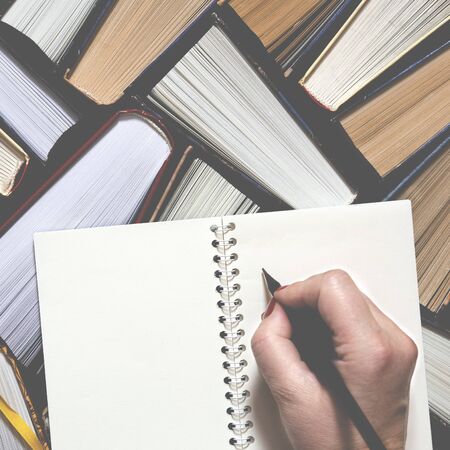 Female hand writing notes to the notebook. Lots of colourful thick open books stand on a dark...