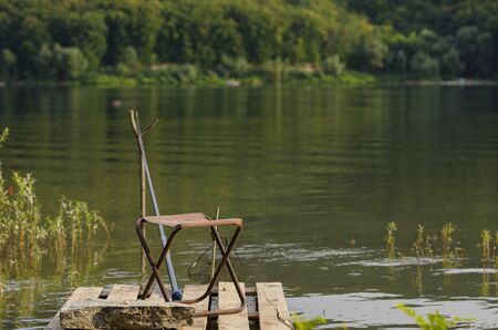 Fisherman's place. A chair, a fishing rod and fish bait rests on a stone against the backdrop of a large river and green forestの写真素材
