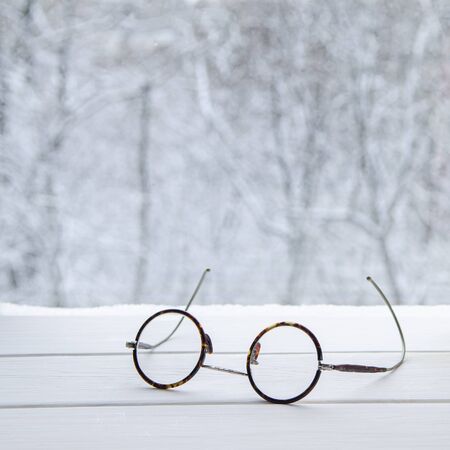 Vintage round reading glasses lie on a white tree window sill against the backdrop of a winter...