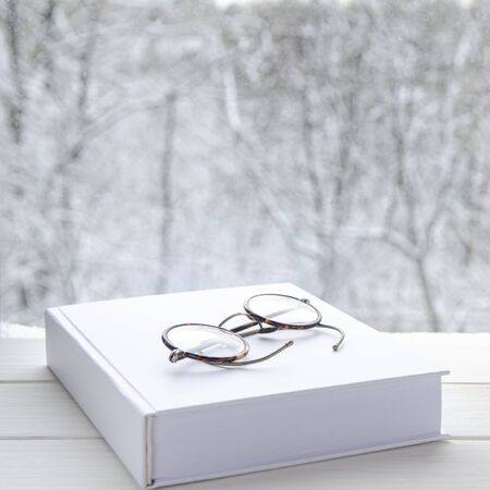 Round vintage reading glasses lie on a white thick notepad against the backdrop of the winter forest