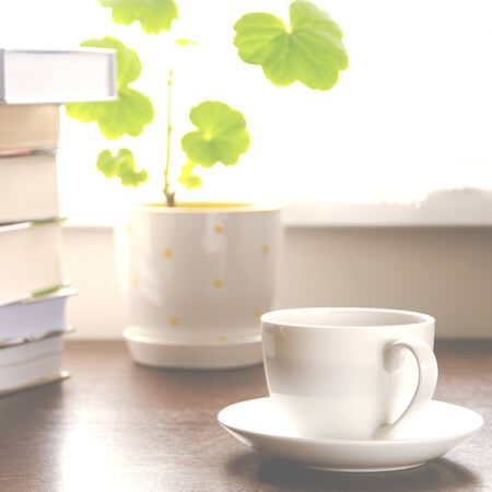 A white cup of coffee stands on a wooden table next to a stack of books and a pot against the background of the window. Close-upの写真素材