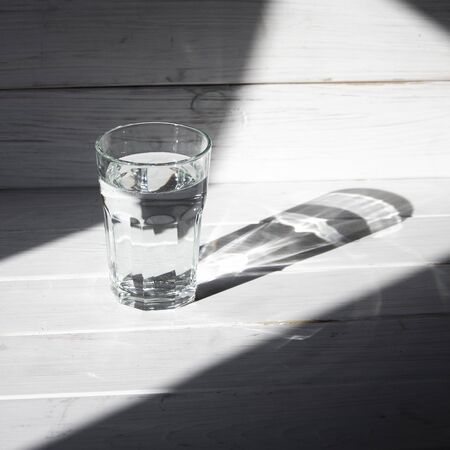 A glass of clean clear water stands on a white wooden background with sharp shadows. shadows
