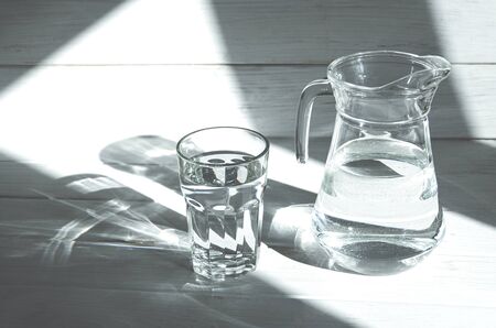 A glass of clean clear water stands on a white wooden background with sharp shadows. shadows