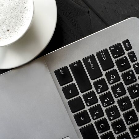 Freelance workplace. A laptop and a cup of coffee with foam stand on a black wooden table. Top view