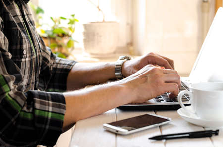 Home office. Freelance workplace. Man's hands on laptop keyboard next to smartphone, pen and cup of coffeeの写真素材
