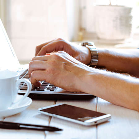 Home office. Freelance workplace. Man's hands on laptop keyboard next to smartphone, pen and cup of...