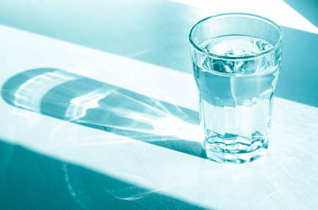 A glass with clean clear water and sharp shadows stands on a white wood table. top view of glass with water on white tableの写真素材