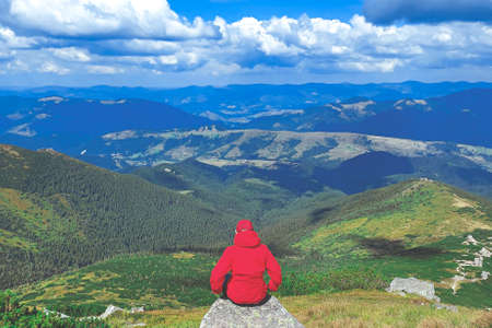 A tourist in a red jacket sits on a stone on top of a mountain against the backdrop of a...