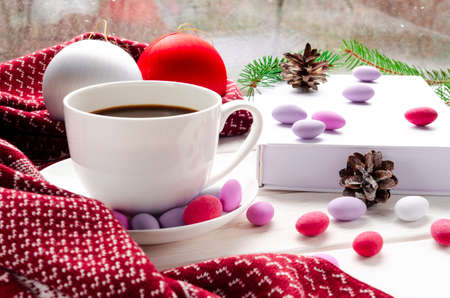 Cozy concept. Cup of coffee with colorful sweets, christmas toys and a warm scarf on a wooden windowsill against the background of a windowの写真素材
